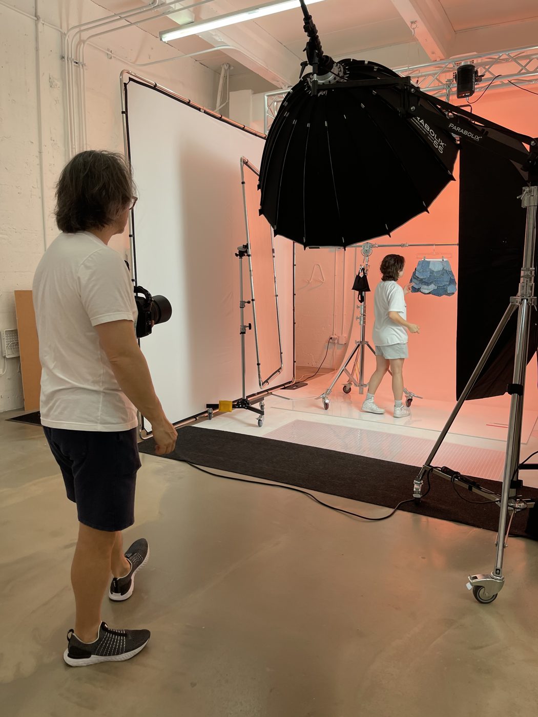 Photographer shooting a model on a coral backdrop in a white cyclorama room with overhead truss